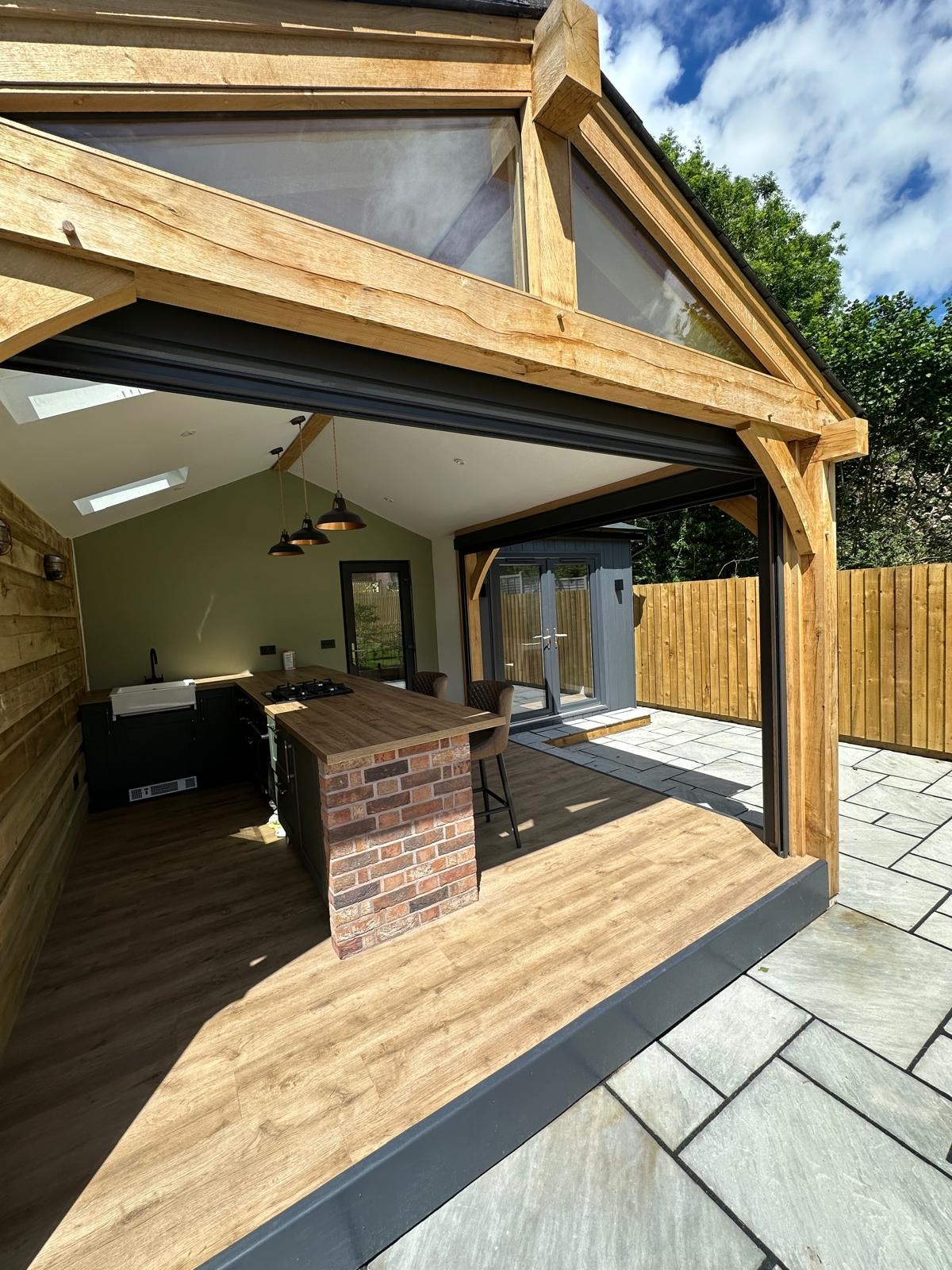 Finished oak-framed garden kitchen with a brick island, pendant lights and open doors to a paved terrace